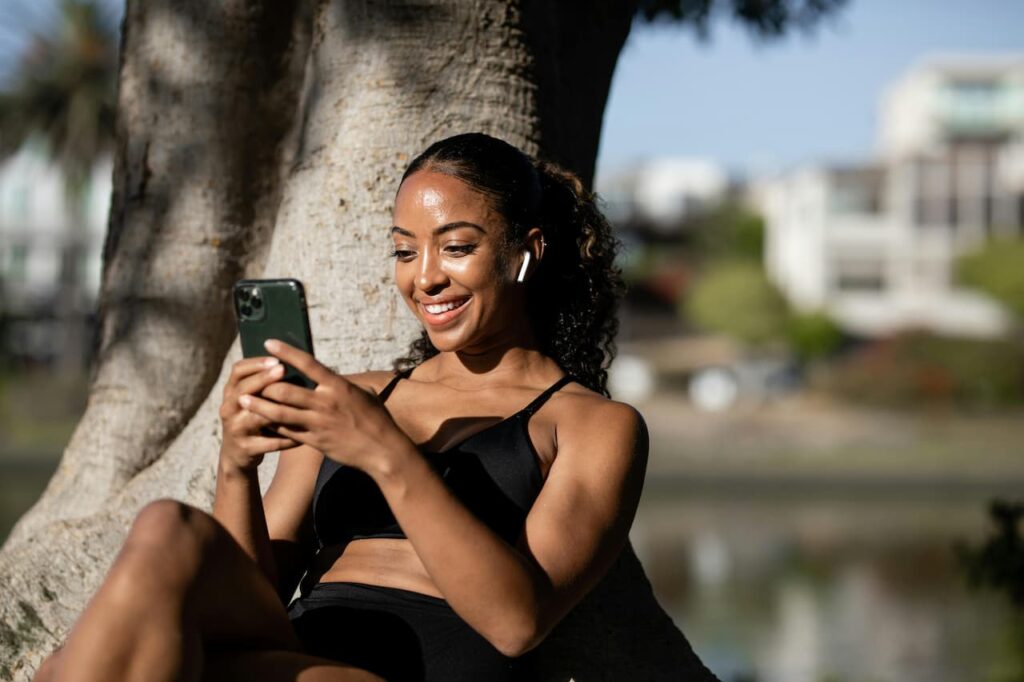 Personne portant des AirPods Apple tout en utilisant un iPhone à l’extérieur, assise contre un arbre.