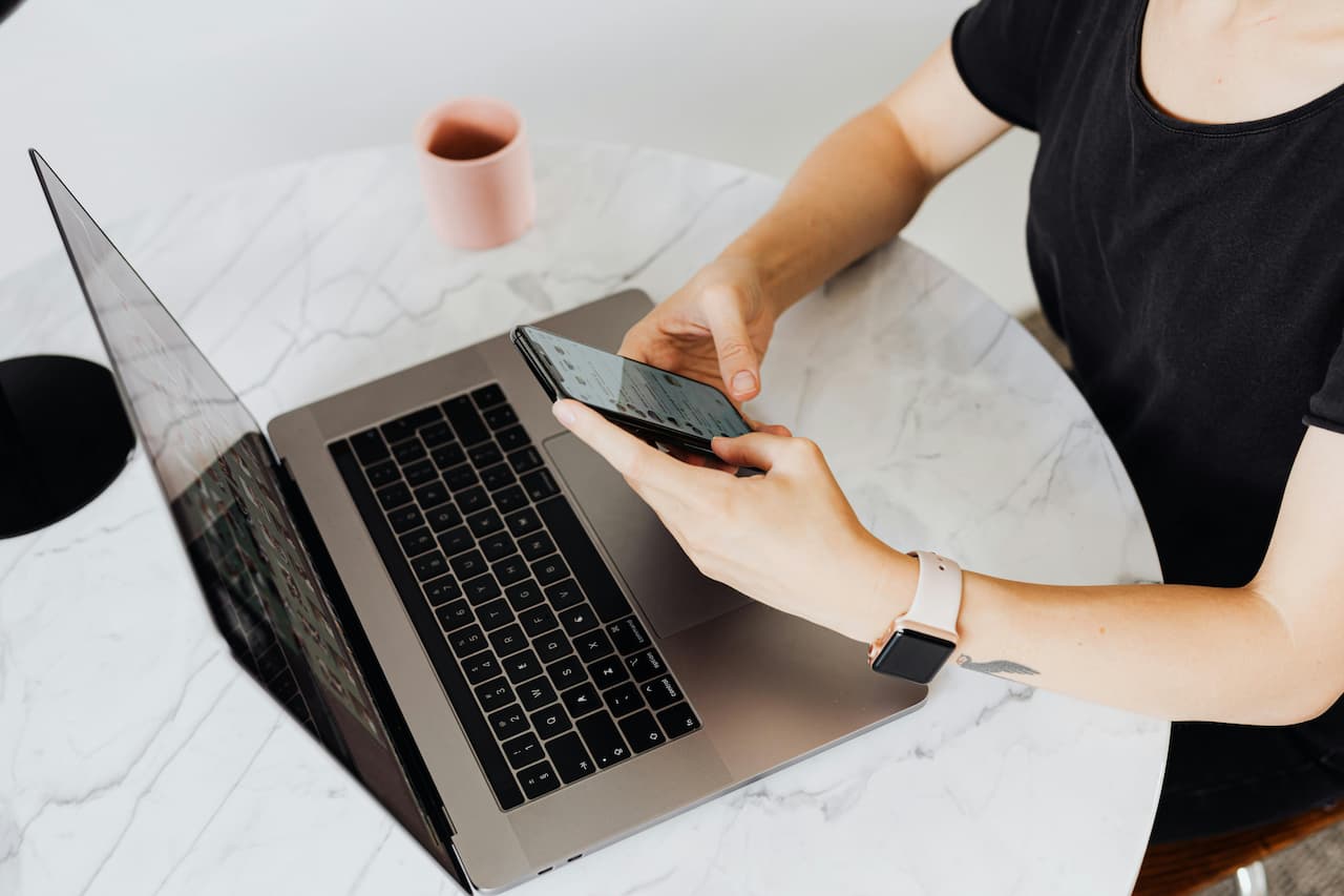 Personne tenant un iPhone devant un MacBook Pro avec une Apple Watch au poignet, connectant ses appareils Apple sur une table en marbre