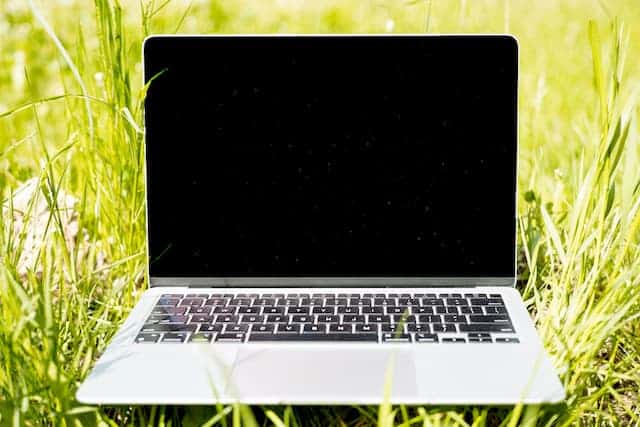 MacBook argenté ouvert posé dans l'herbe verte en plein soleil, écran noir, illustrant les risques de surchauffe en extérieur l'été