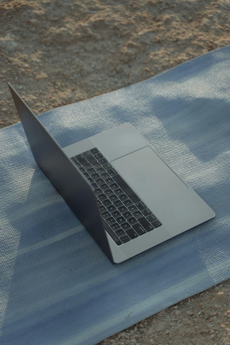 MacBook argenté posé à plat sur un tapis bleu au bord du sable de plage, exposé à la chaleur et aux risques liés au sable et à l'humidité
