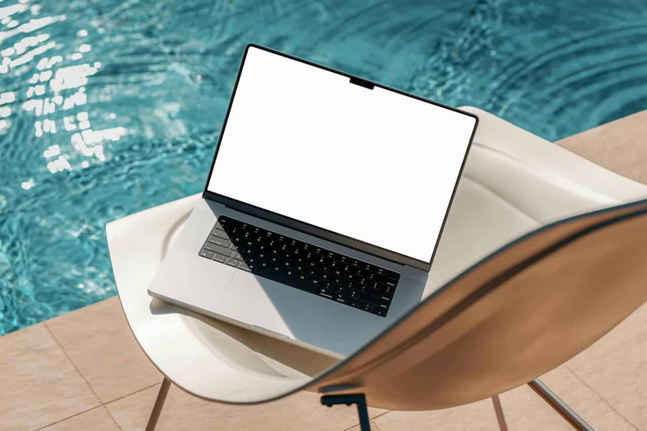 MacBook Pro ouvert posé sur une chaise longue blanche au bord d'une piscine en plein soleil, risque de surchauffe et d'humidité