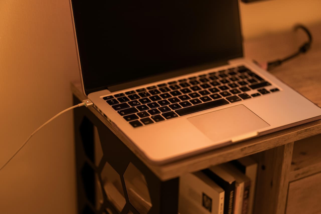 MacBook branché en permanence avec son chargeur MagSafe allumé, posé sur une étagère en bois dans une lumière orangée